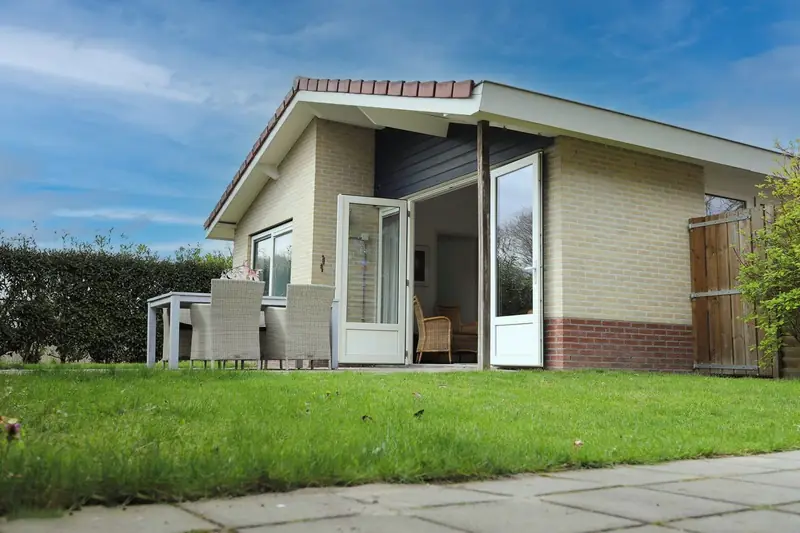 De achterkant van huisje&nbsp;2, met open terrasdeuren, de tuintafel met stoelen en het gazon.