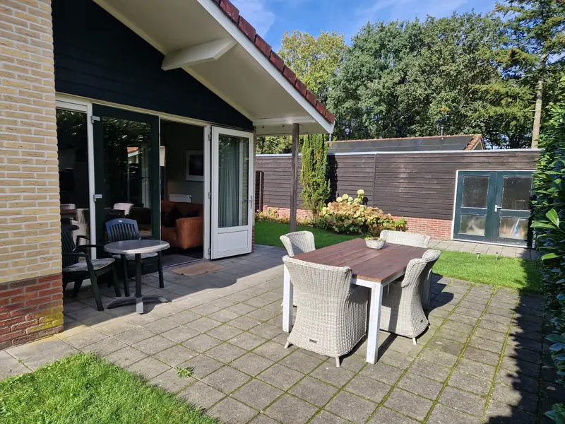 The terrace of cottage&nbsp;2: a wooden table with four wicker chairs, a bistro table by the open French doors, and the shed beyond the lawn.